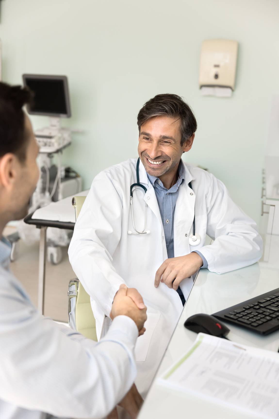 Ein lächelnder Arzt mit weißem Kittel und Stethoskop sitzt an einem Schreibtisch und schüttelt einem Patienten in einem medizinischen Büro die Hand. Ein Computer, Papierkram und medizinische Geräte sind im Hintergrund zu sehen.