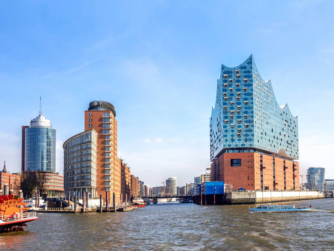 ©pure-life-picturesstock/adobe.stock.com Eine Stadtlandschaft am Wasser mit modernen und historischen Gebäuden, darunter die gläserne Elbphilharmonie in Hamburg, Deutschland, unter einem klaren blauen Himmel, mit Booten auf dem Fluss im Vordergrund.