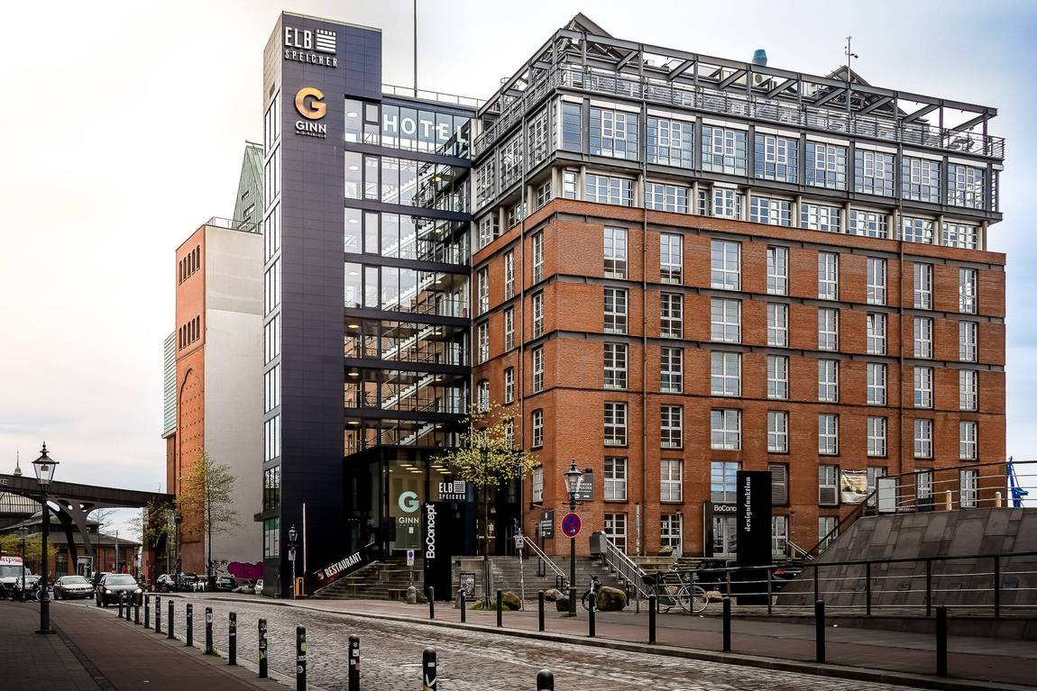 Ein modernes, mehrstöckiges Gebäude aus Ziegeln und Glas mit der Aufschrift GINN Hotel und Elbarkaden steht an einer Stadtstraße mit Kopfsteinpflaster, Pollern und nahe gelegenen Laternenpfählen.