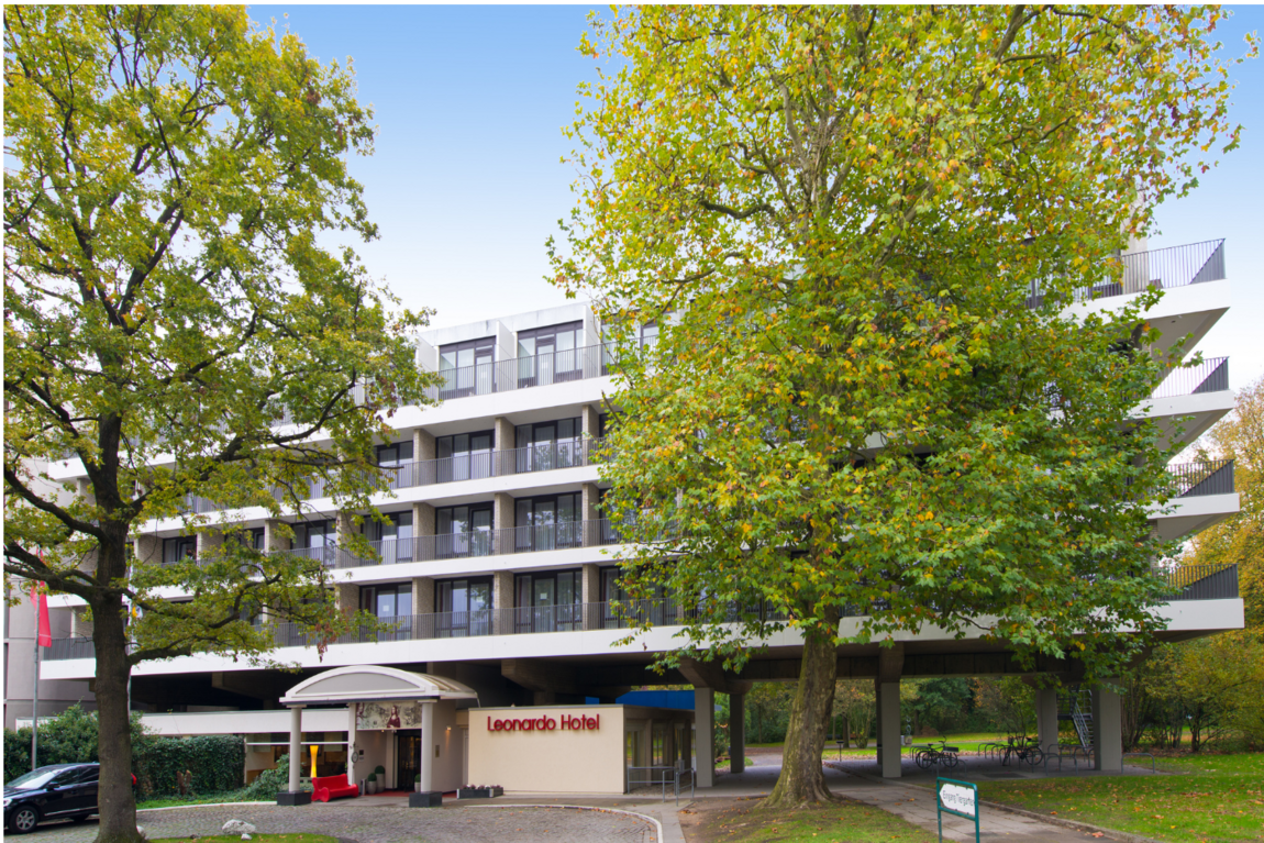 ©Leonardo Hotels Ein modernes, mehrstöckiges Leonardo-Hotel mit Balkonen wird teilweise von großen grünen Bäumen verdeckt. Der Hoteleingang hat ein kleines Vordach, und ein Auto ist in der Nähe der geschwungenen Einfahrt geparkt. Der Himmel ist klar und blau.