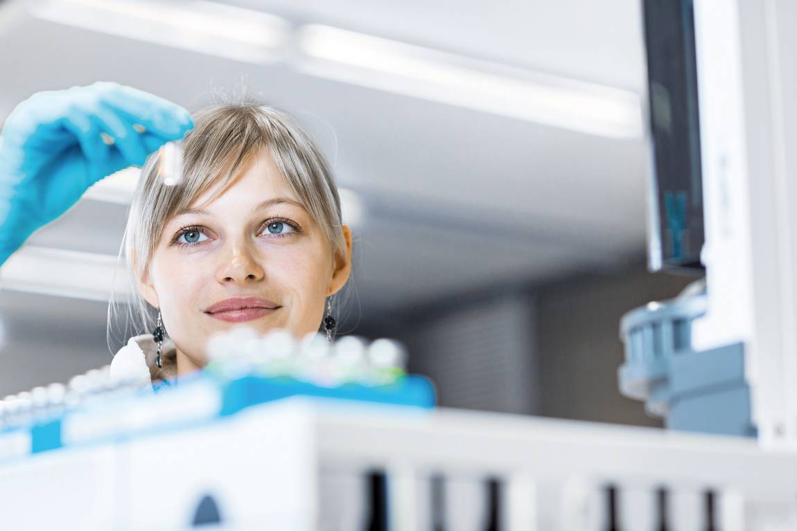 ©istock.com/ViktorCap Ein Wissenschaftler mit blauen Handschuhen untersucht ein kleines Fläschchen in einer Laborumgebung, umgeben von wissenschaftlichen Geräten und Fläschchen.
