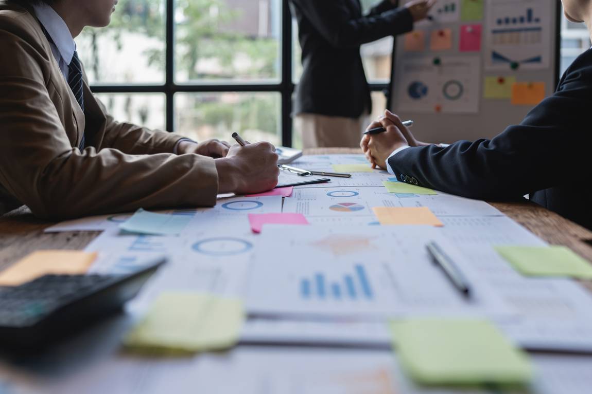 Drei Personen in Geschäftskleidung befinden sich in einem Besprechungsraum mit Diagrammen, Schaubildern und Haftnotizen auf einem Tisch. Eine Person präsentiert an einer Tafel, während zwei andere sitzen und Notizen machen. Helles Fenster im Hintergrund.