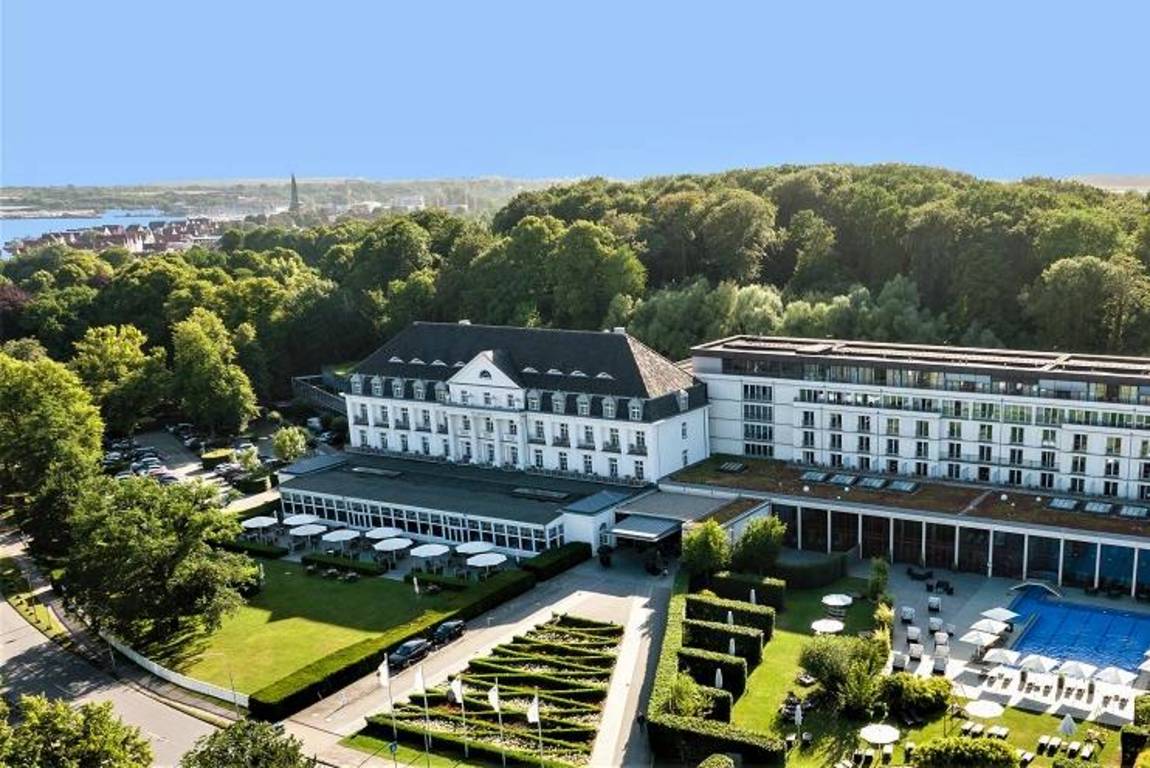 Luftaufnahme eines großen, eleganten Hotels, umgeben von üppigen grünen Bäumen, mit Gärten, Sitzgelegenheiten im Freien und einem blauen Swimmingpool; im Hintergrund sind eine Stadt und Wasser unter einem klaren blauen Himmel zu sehen.